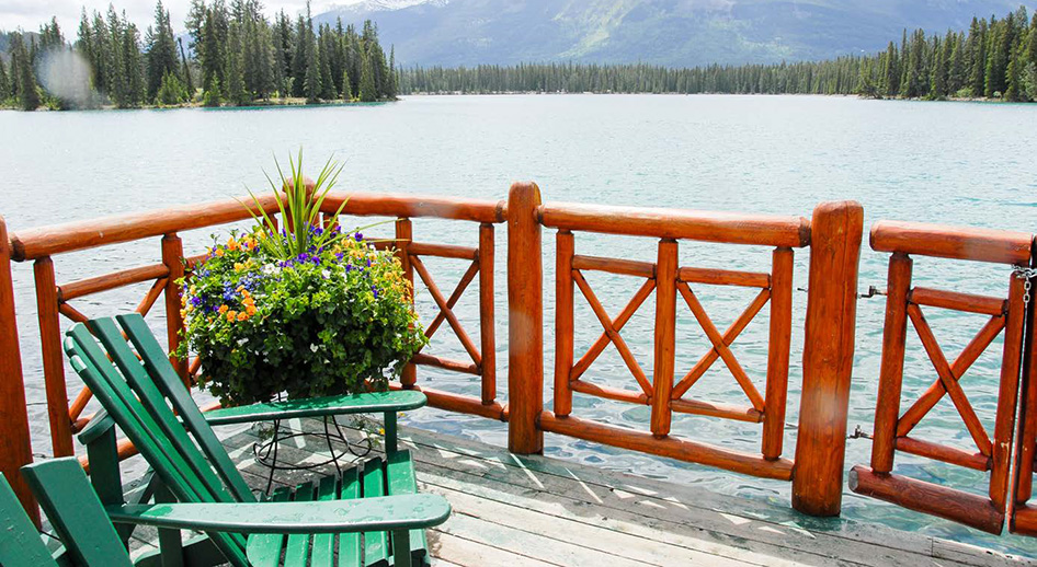 Lake side patio with green adirondack chairs. In the background the far shore of the lack is thick with evergreen trees at the foot of distant mountains. 