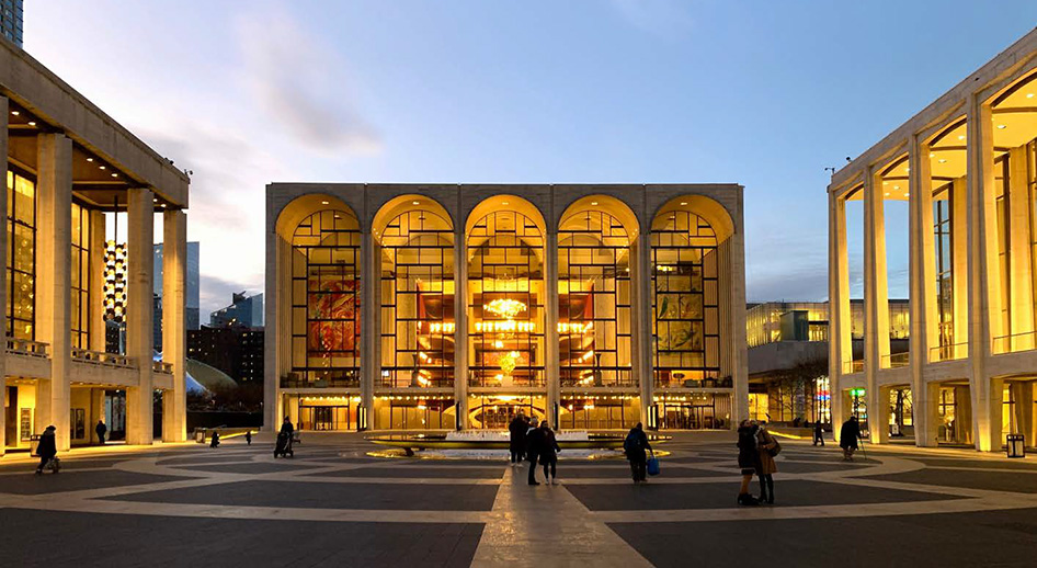 Warm light illuminates the New York Metropolitan Opera House under a clear evening sky.
