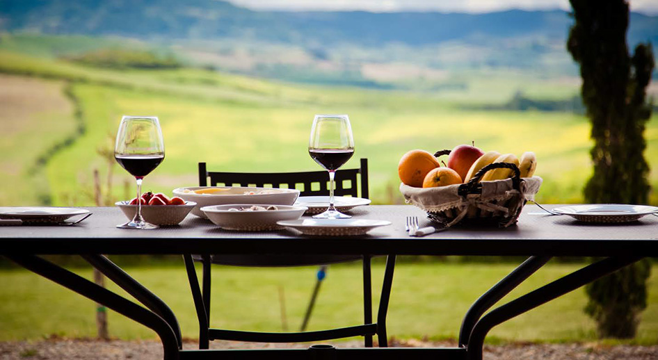 Outdoor table set for dinner with a basket of fruit and glasses of read wine. A scenic view of the Italian country side is in the background.